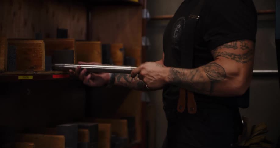 A muscular, tattooed worker inspects and selects various metal tools and parts from a storage shelf in his workshop
