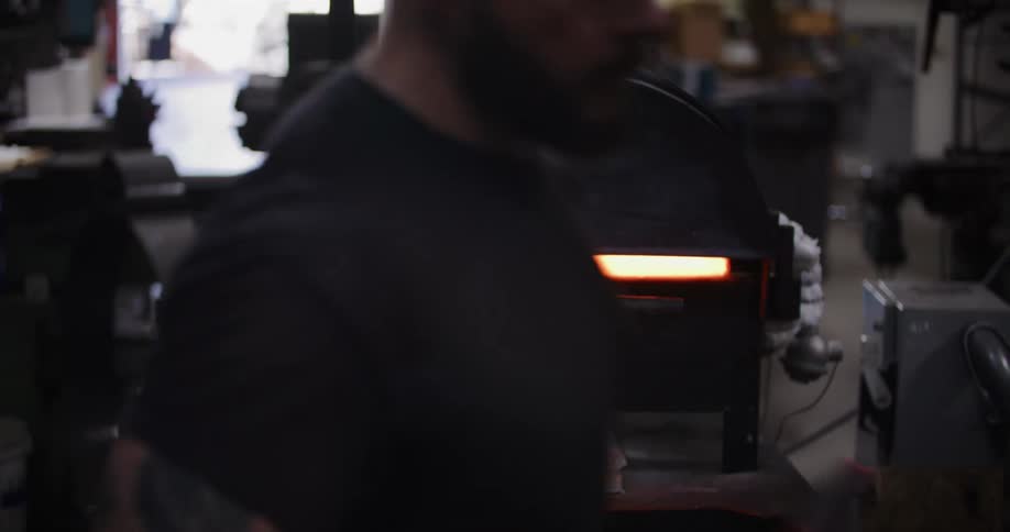 A metalworker removes a piece of red-hot steel from a furnace in a dark, industrial workshop