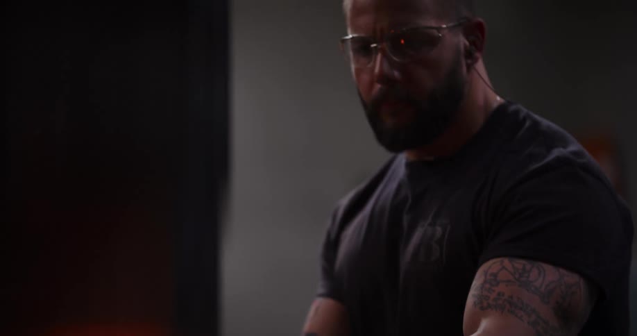 A muscular blacksmith with tattoos carefully removes a glowing hot piece of metal from a fiery forge