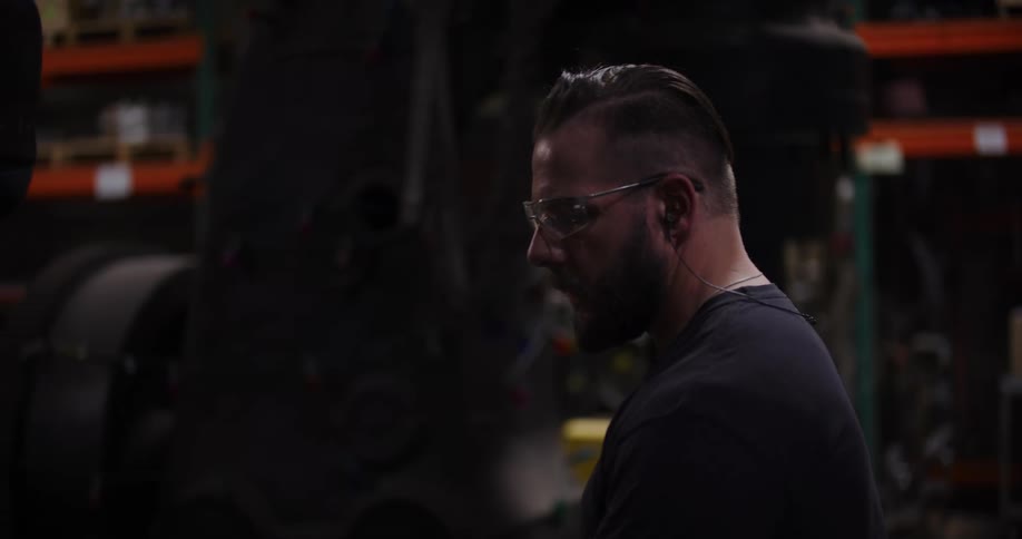 Side profile portrait of a focused male factory worker wearing safety glasses in a dimly lit industrial warehouse