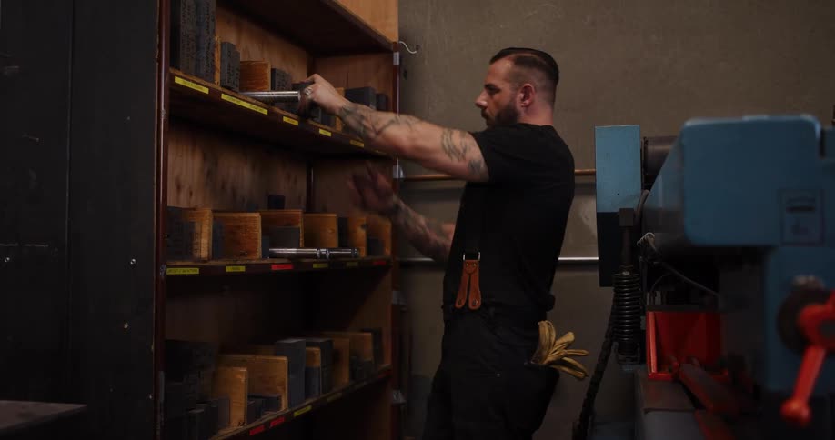 A tattooed, muscular male worker carefully selects a metal die from a storage shelf in a fabrication workshop