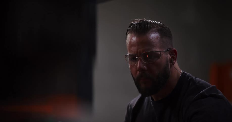An intense close-up shot shows a rugged, tattooed blacksmith with a beard and safety glasses concentrating on his craft in a dimly lit forge