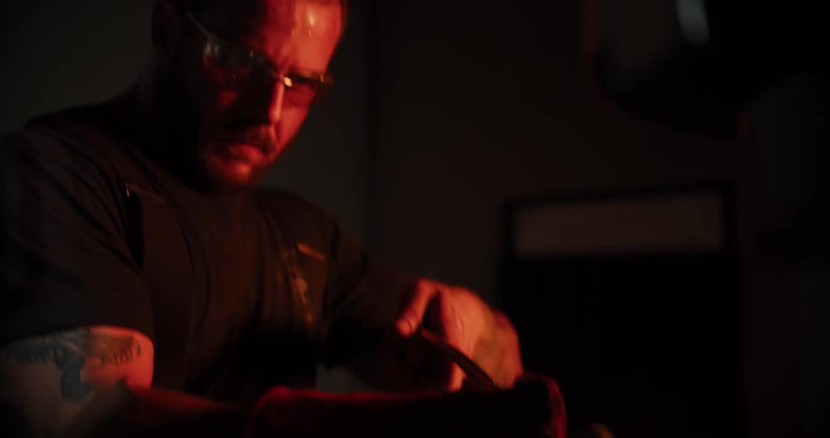 A focused blacksmith wearing safety glasses and protective gloves carefully handles a glowing red-hot metal billet with tongs in his dark forge