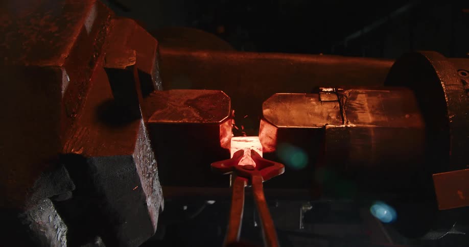 A power hammer forges a red-hot billet of steel held by tongs in a dark industrial workshop