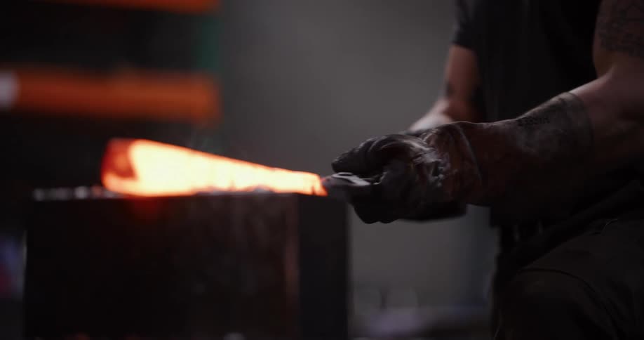 A muscular blacksmith in slow motion uses a power hammer to forge a glowing hot piece of metal, creating sparks