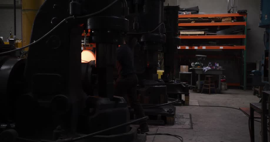 A blacksmith uses a large industrial power hammer to forge a piece of glowing hot metal in his workshop