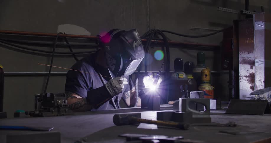 A skilled metalworker in a protective mask carefully welds metal components on a workbench in a dimly lit industrial workshop