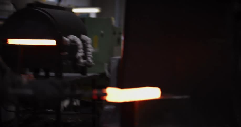 A blacksmith removes a glowing hot piece of steel from a forge and shapes it with a power hammer, sending sparks flying