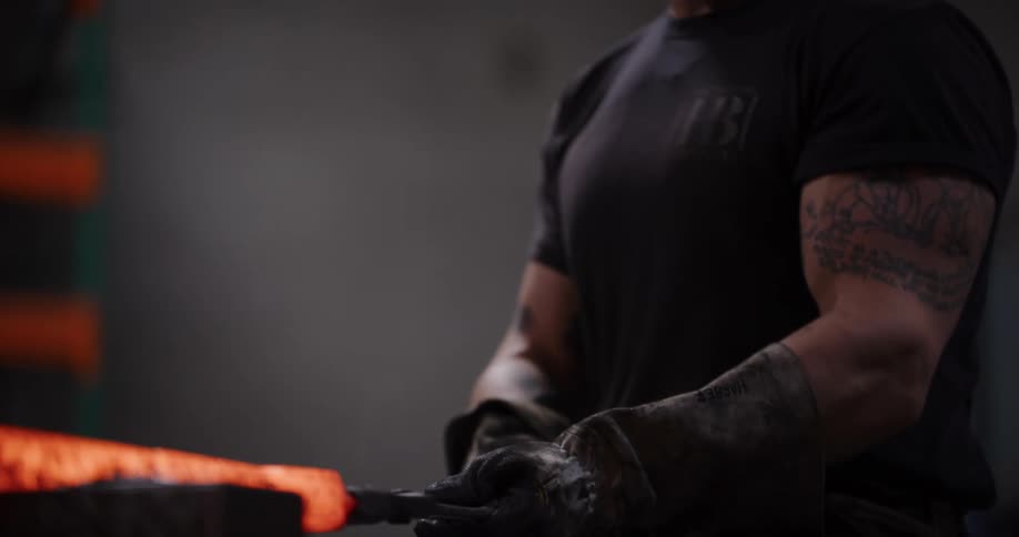 A muscular blacksmith with tattoos uses a power hammer to forge a piece of glowing red-hot metal in his workshop