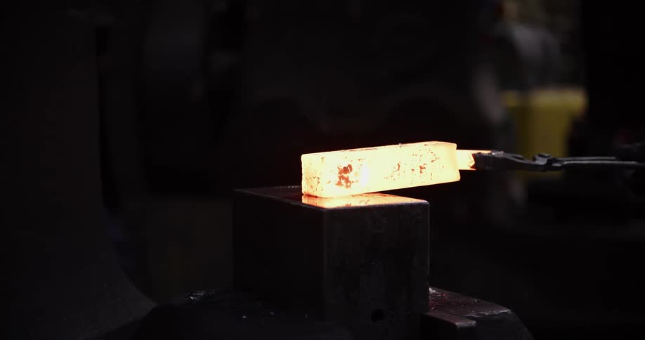 A blacksmith uses tongs to pull a glowing hot metal billet from a forge and shapes it under a powerful mechanical press in dramatic slow motion