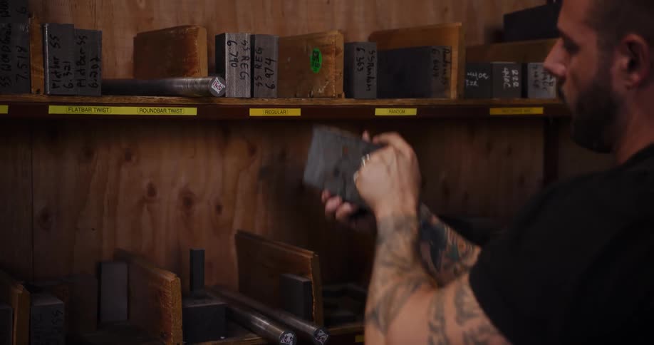A tattooed craftsman carefully inspects and selects pieces of raw metal stock from a workshop shelf