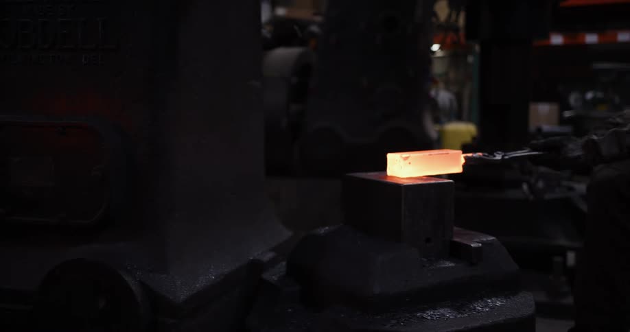 A skilled blacksmith pulls a glowing hot piece of metal from a forge and uses a massive power hammer to shape it, sending sparks flying in a dramatic workshop scene