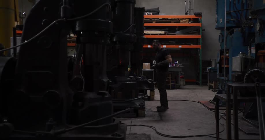 A muscular blacksmith walks through his workshop to operate a large industrial power hammer