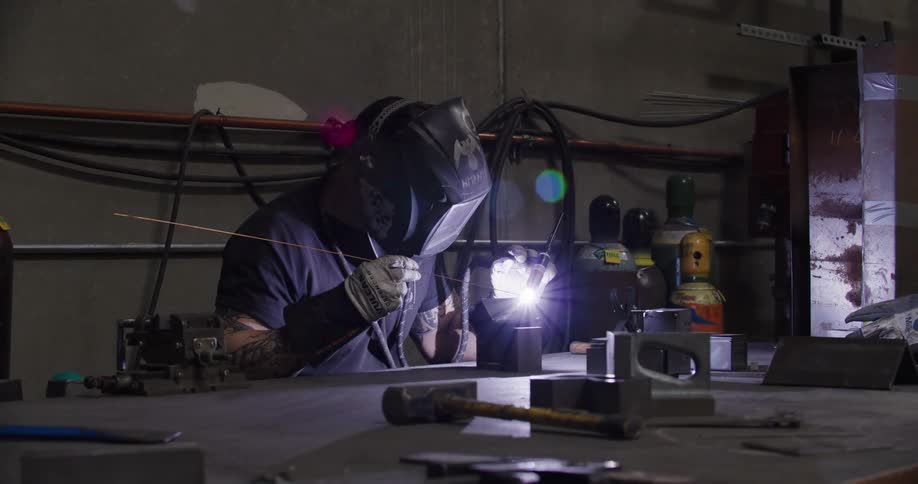 A welder lowers his protective helmet and begins to TIG weld a piece of metal on his workbench in a dark workshop