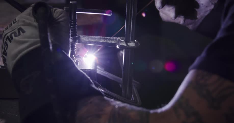 A tattooed craftsman meticulously TIG welds a metal joint clamped in place, creating an intense, bright light in a dark workshop