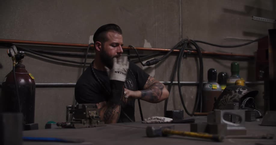 A tattooed welder prepares for his work by putting on protective gloves and his welding helmet in a dimly lit workshop