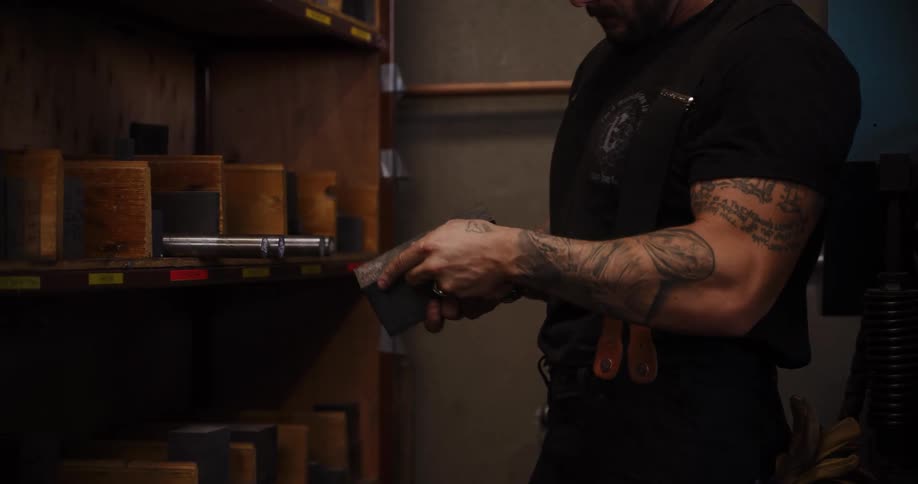 A tattooed craftsman in his workshop carefully selects and inspects a metal die from a wooden shelf in slow motion
