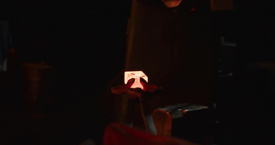 A blacksmith takes a glowing piece of metal from a furnace and shapes it with a power hammer in a dark workshop