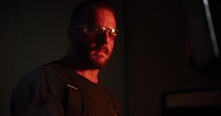 A skilled blacksmith in safety glasses carefully inspects a glowing red-hot metal blade in his dark forge