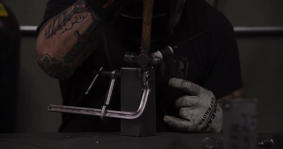 A welder with tattooed arms uses a TIG torch to meticulously weld a metal part in a slow-motion close-up