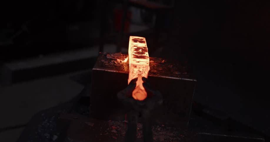A blacksmith uses tongs to pull a glowing hot piece of metal from a forge and shapes it with a power hammer, sending sparks flying