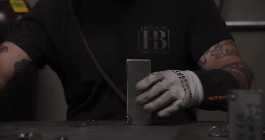 A tattooed welder in his workshop removes a C-clamp and uses a hammer to shape a piece of metal