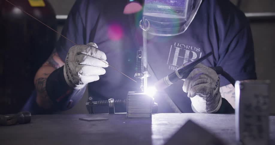 A welder in a protective mask uses a TIG torch to carefully weld a metal piece in a workshop, creating a bright purple arc