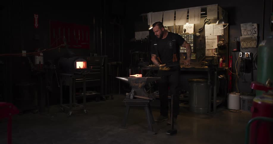 A blacksmith pulls a glowing hot piece of metal from a forge and places it on an anvil in his workshop