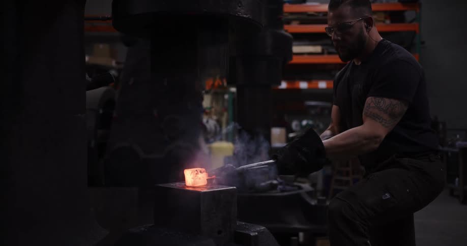 A blacksmith carefully uses a large power hammer to shape a piece of glowing hot metal in his workshop
