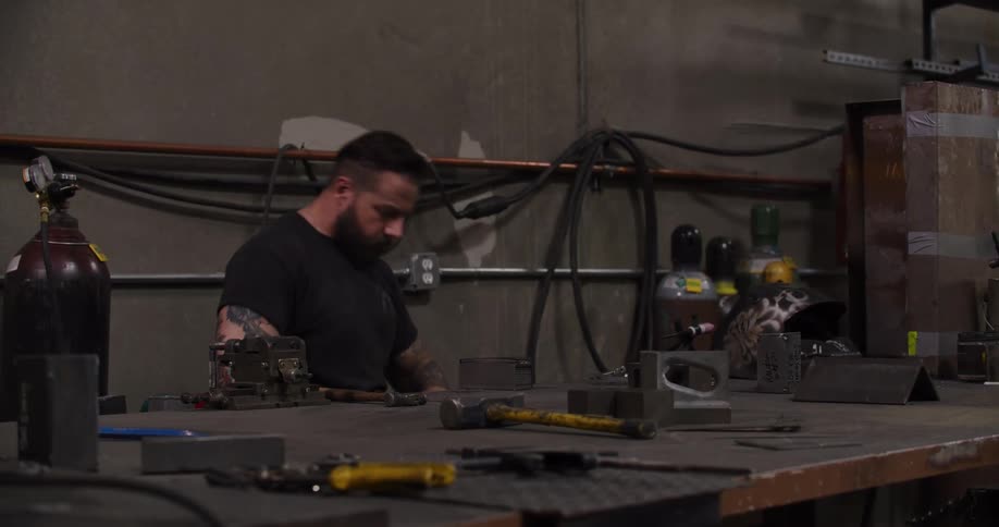 A tattooed craftsman carefully measures a block of metal with calipers at his workbench in a metal shop