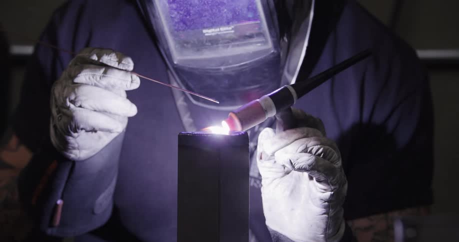 A skilled welder in a protective mask carefully uses a TIG torch to weld a metal post, creating a bright purple arc