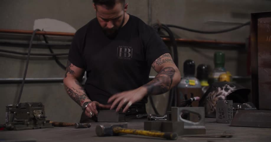 A tattooed craftsman with a beard sands and clamps a stack of metal plates on his workbench in a fabrication shop