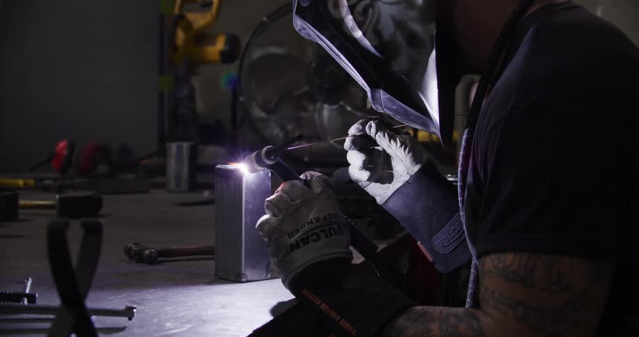 A welder with tattooed arms wears a protective mask while TIG welding a metal object in a dark workshop, creating a bright purple arc and sparks