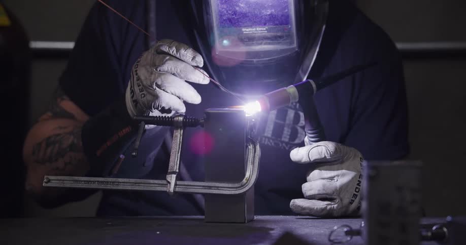 A welder carefully TIG welds a metal piece held in a clamp in a dimly lit workshop