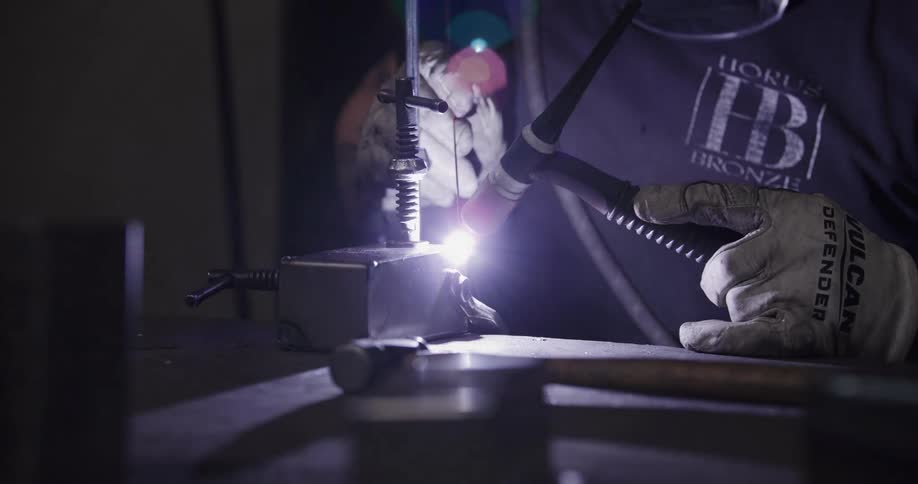 A close-up shot of a welder meticulously TIG welding a metal piece, with a brilliant flash of light illuminating the dark workshop
