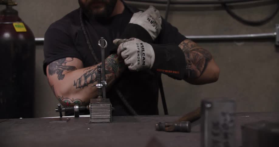 A tattooed metalworker puts on protective gloves and lowers his helmet to begin welding in his workshop