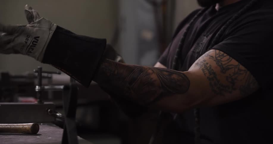 A muscular, tattooed welder carefully puts on his protective gloves and sleeves before beginning to weld in his workshop