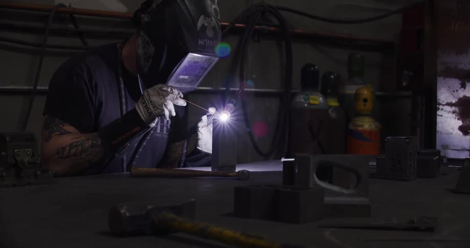 A tattooed welder in a protective mask sits at a workbench in a dark shop, skillfully creating a bright arc of light to fuse metal