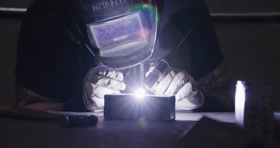 A welder wearing a protective mask and gloves carefully performs TIG welding on a metal block in a dark workshop