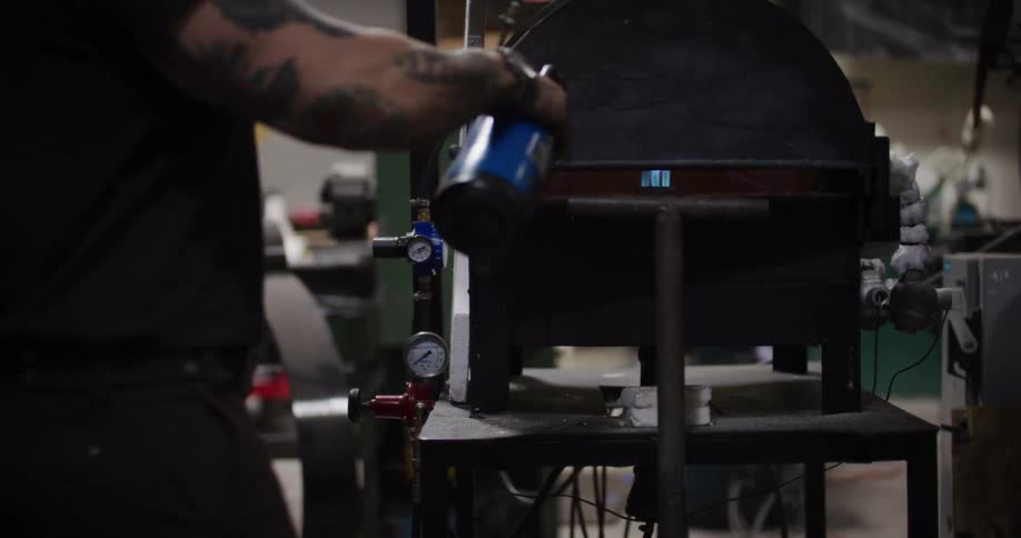 A craftsman with tattooed arms uses a blowtorch on a furnace before closing the lid in a workshop