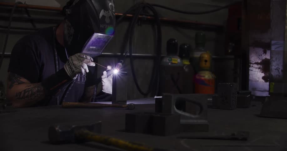 A welder with tattoos wears a protective helmet while carefully working on a metal piece in a dark workshop