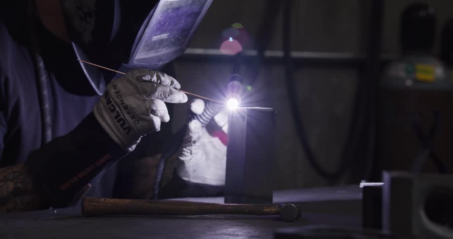 A skilled metalworker in a protective mask carefully TIG welds a piece of metal in a dimly lit workshop