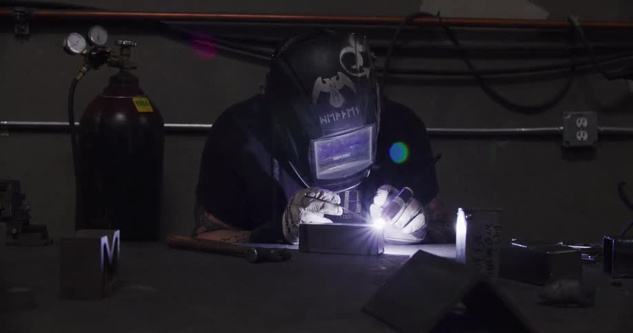 A welder lowers his protective helmet and begins TIG welding a metal part on a workbench in a dark workshop