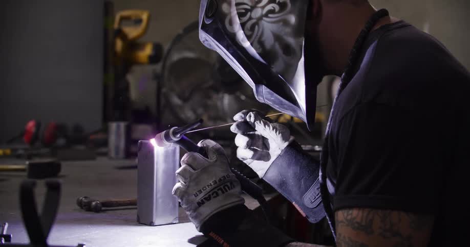 A bearded metalworker puts on a custom welding helmet and carefully begins to TIG weld a piece of steel in his workshop