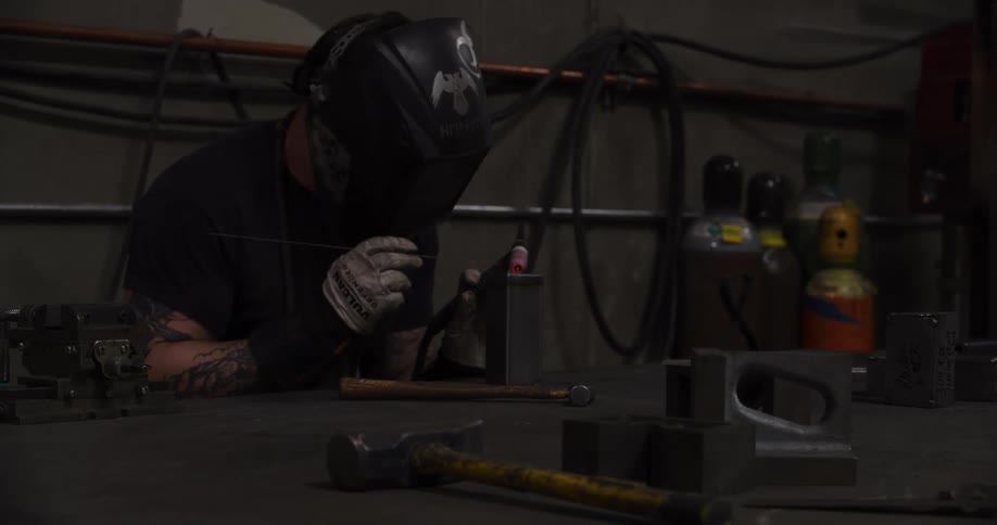 A skilled welder with tattoos meticulously joins metal pieces on a workbench in a dimly lit industrial workshop