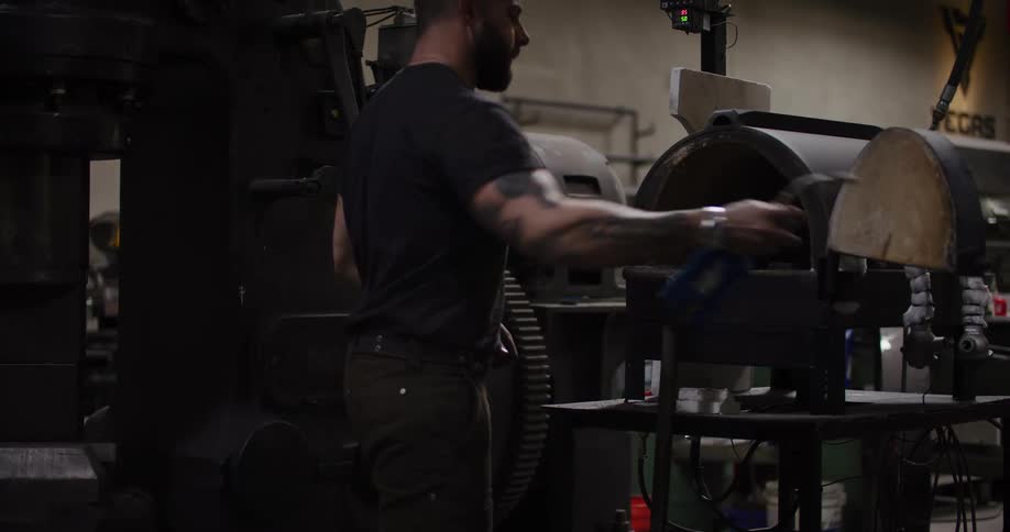 A muscular, tattooed blacksmith lights a furnace with a propane torch in his industrial workshop