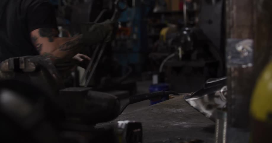 A tattooed man prepares his welding equipment at a workbench in a dim industrial workshop