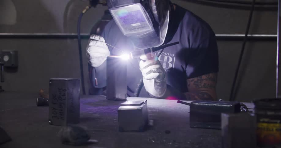 A skilled welder with tattoos lowers his protective helmet and carefully TIG welds a metal component on his workbench