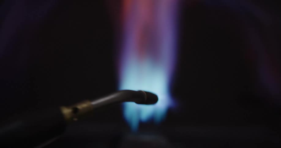 A blowtorch ignites a powerful gas burner, revealing a vibrant blue, purple, and orange flame in slow motion against a dark background
