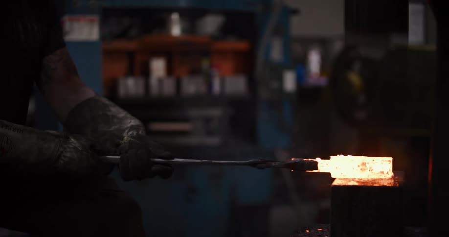 A blacksmith uses an industrial power press to forge a glowing hot piece of metal in a dark workshop